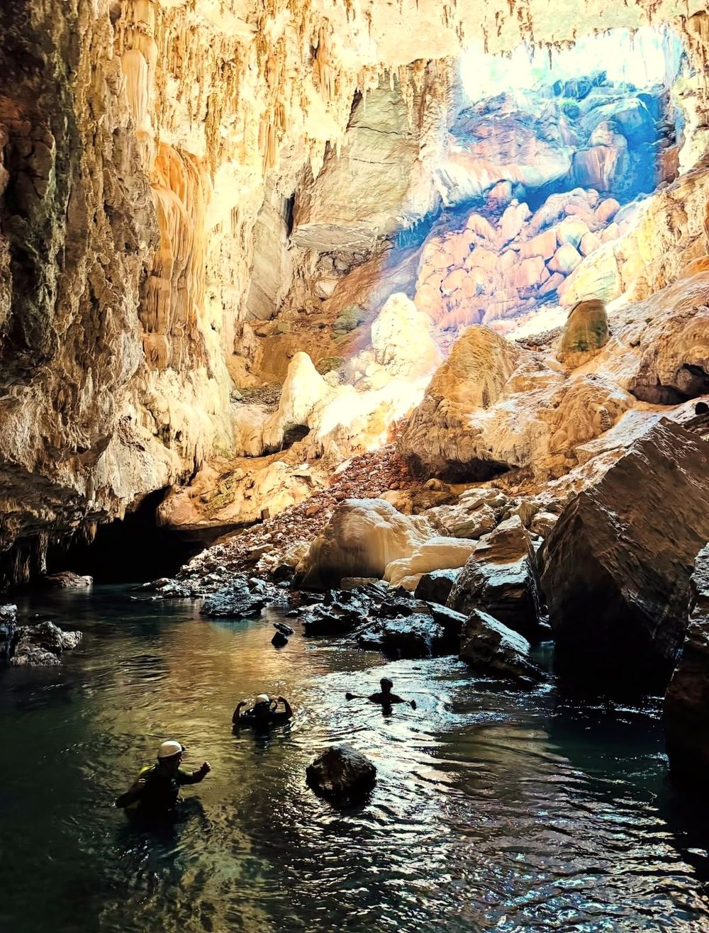 Pessoas nadando num rio dentro da caverna, com luz do dia entrando por uma abertura no alto