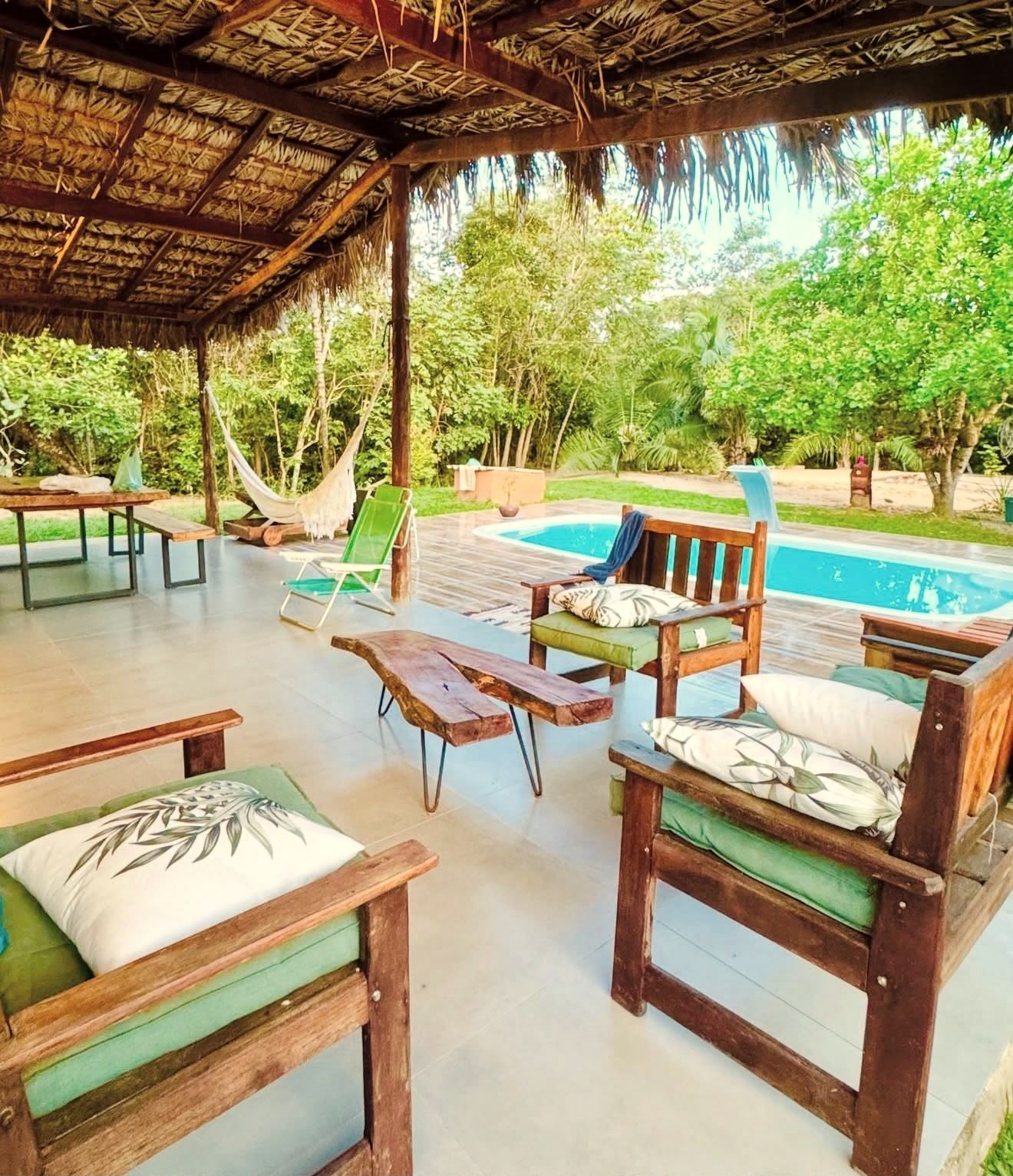 Área coberta com poltronas de madeira e mesa de tronco, de frente para a piscina e o gramado